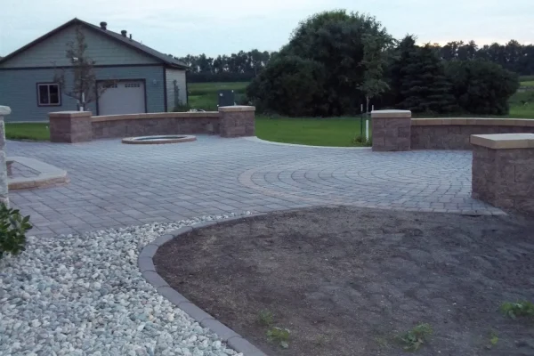 A paved patio area with a circular fire pit and surrounding stone walls, near a garage and landscaped yard.