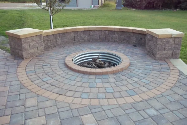 Circular stone patio with a fire pit and a low curved wall, bordered by grassy lawn and a building in the background.