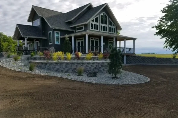 A modern two-story house with large windows, a wraparound porch, and a landscaped yard showcasing expert hardscaping & landscaping, including gravel paths, shrubs, a stone retaining wall, and a spacious dirt driveway.