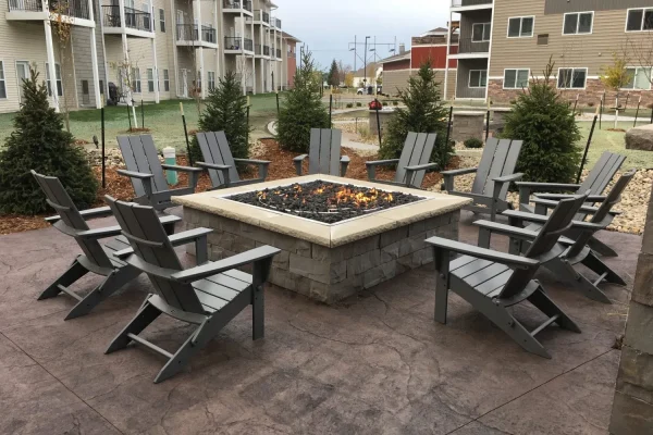 A square outdoor fire pit with black stones is the centerpiece of this inviting Outdoor Living space, surrounded by grey Adirondack chairs on a patio, with apartment buildings and evergreen trees in the background.