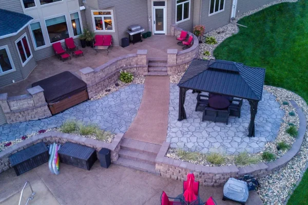 Backyard patio area with a hot tub, seating, a grill, and a gazebo, surrounded by plants and a stone retaining wall.