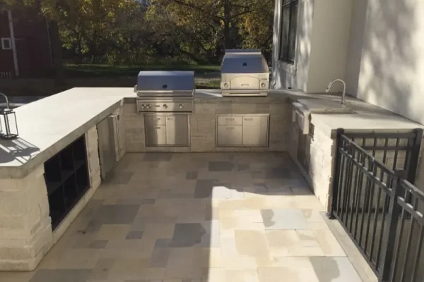 Outdoor Living shines in this kitchen with stone counters, built-in stainless steel grills, storage cabinets, a sink, and open shelving, all set on a tiled patio surrounded by trees and sunlight.