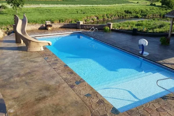 A stunning rectangular outdoor swimming pool with a slide and basketball hoop, surrounded by stone-patterned decking and grassy fields—perfect for embracing outdoor living. The sunlit pool area is empty, offering a serene escape.