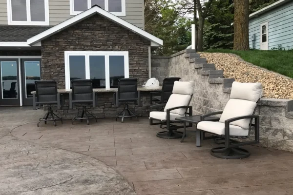 Patio area with stunning hardscaping stone walls, several cushioned swivel chairs, a row of barstools at an outdoor counter, and a house in the background with stone siding and large windows—perfect for seamless landscaping style.